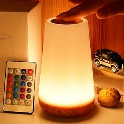 A warm white LED night light lamp with a wooden base, shown glowing on a tabletop with a hand touching the top. A matching remote control and product box are visible beside it, along with a small toy car and a walnut in a room setting.